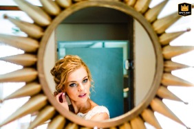 A bride in Playa Mujeres, Cancun, prepares for her wedding. She puts on earrings, checking her reflection in a sun shaped mirror. The photo captures a candid moment of bridal preparation.